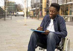 Young man in a wheelchair writing a letter outside.